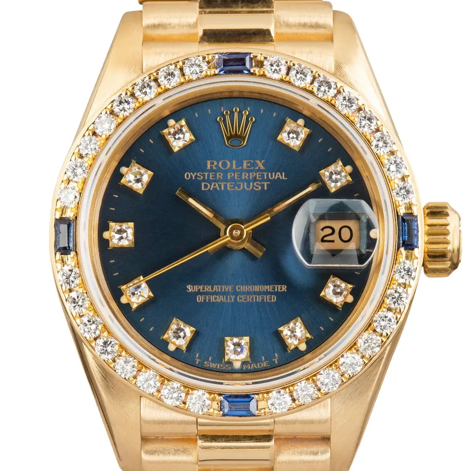 Rolex Lady-Datejust 26 69088 with a blue vignette dial with diamond hour markers, bezel set with diamonds and sapphires and yellow gold President bracelet, a pre-owned example, studio close-up of the case
